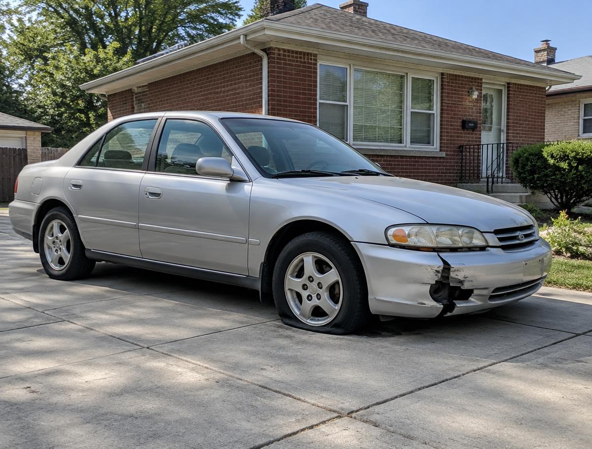 Older silver sedan with bumper damage and a flat tire in a residential driveway